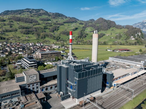 Industrieanlage mit rot-weissem Schornstein, umgeben von grünen Hügeln und einem kleinen Dorf unter klarem blauen Himmel.