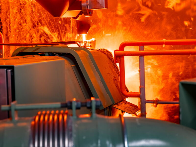 Industrial furnace with glowing molten material and bright sparks surrounded by machinery and orange protective railings.